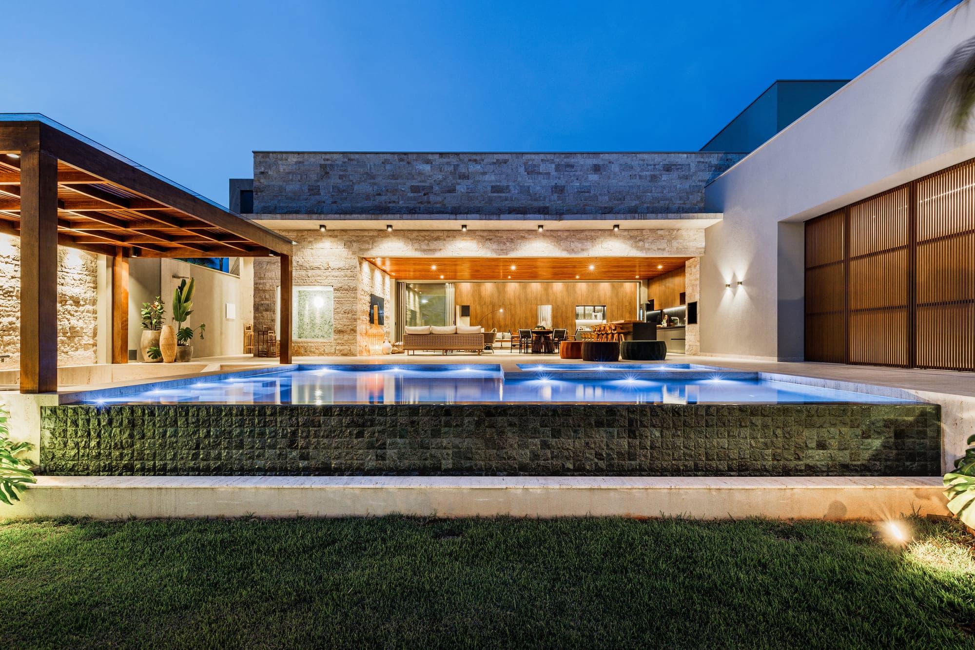 Casa vista do jardim ao crepúsculo, com piscina iluminada em azul e interior aceso