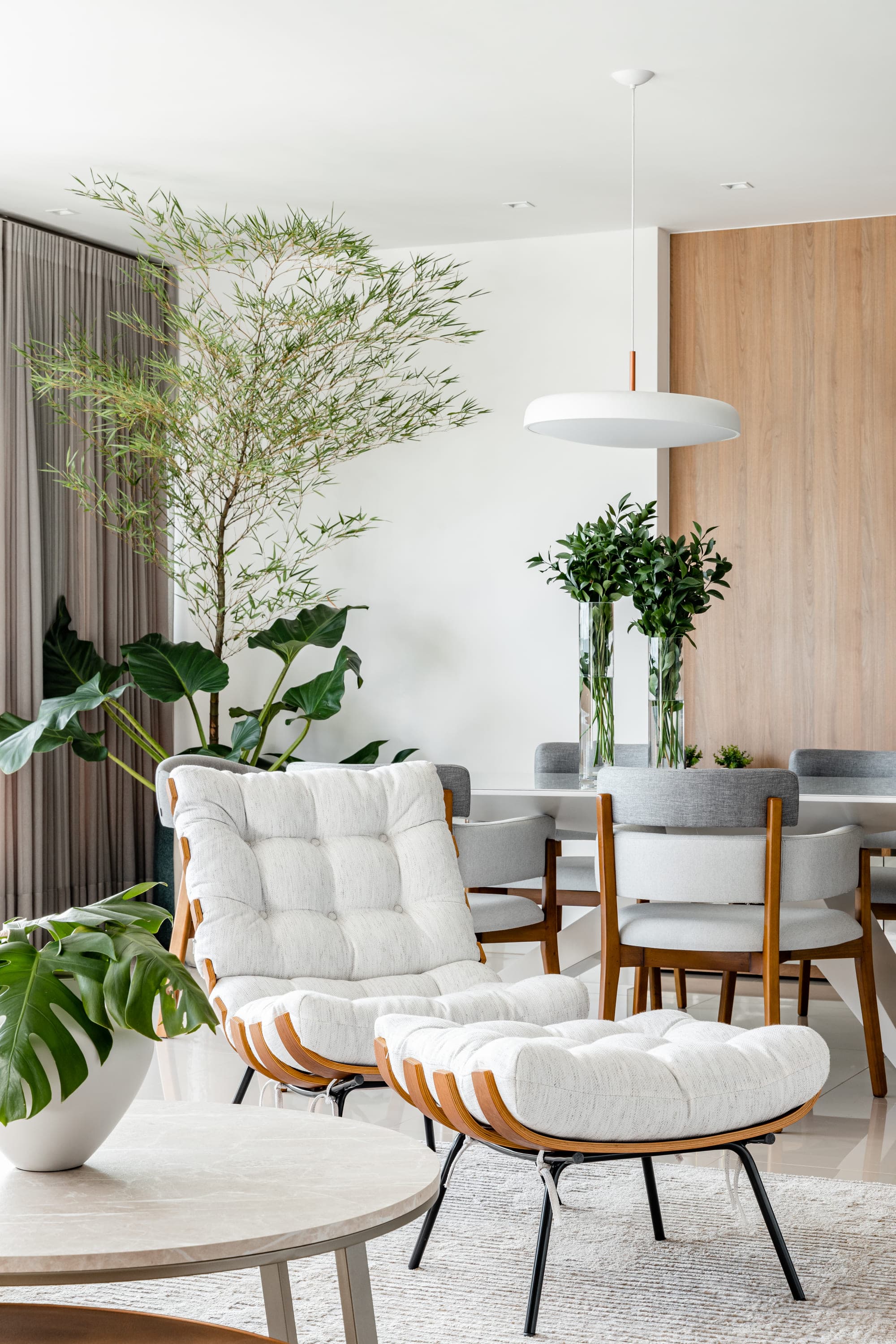 Sala de jantar com mesa de madeira, poltronas brancas estofadas e plantas junto à janela