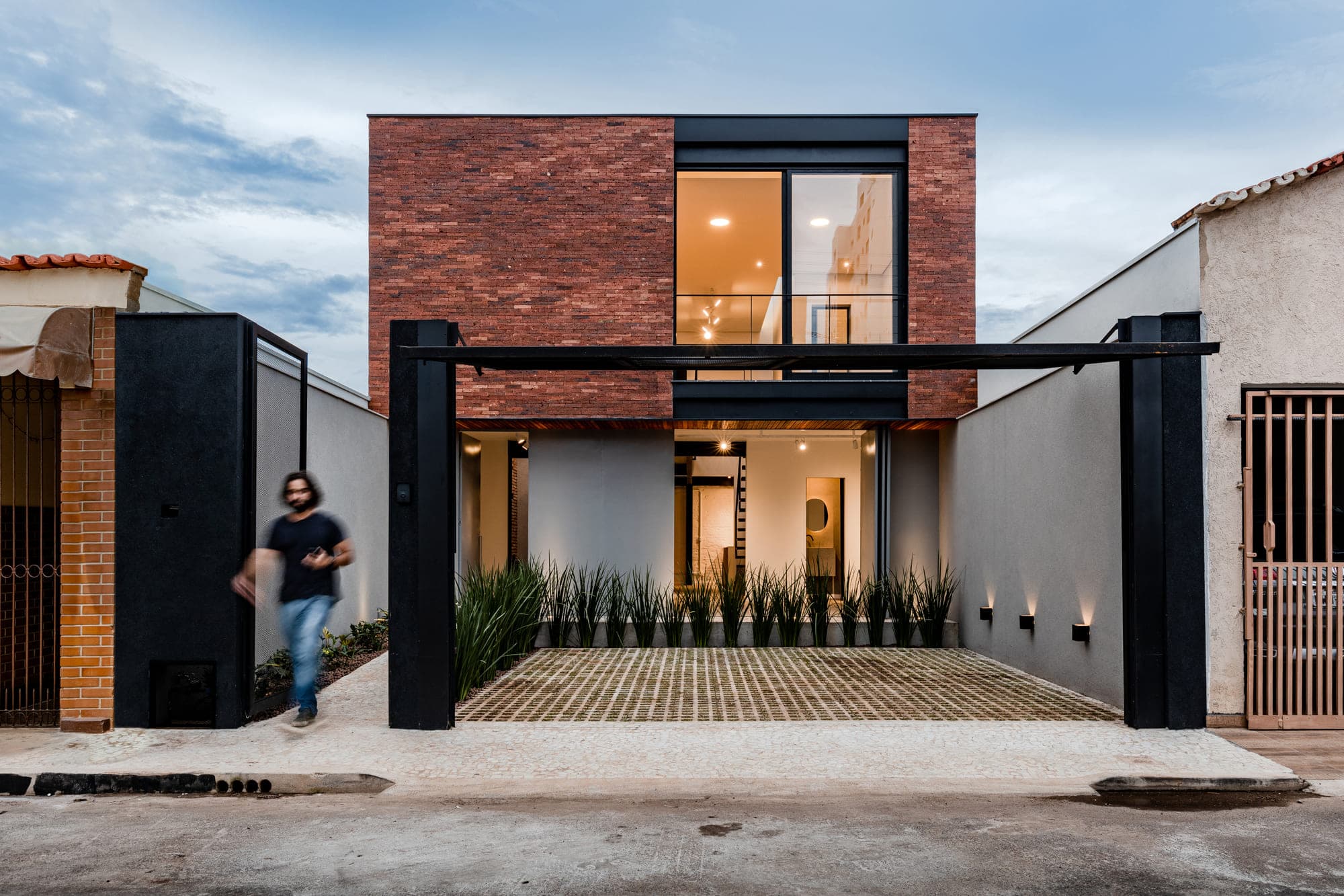 Fachada de casa em tijolo aparente com estrutura metálica preta, fotografada no fim do dia com o interior iluminado