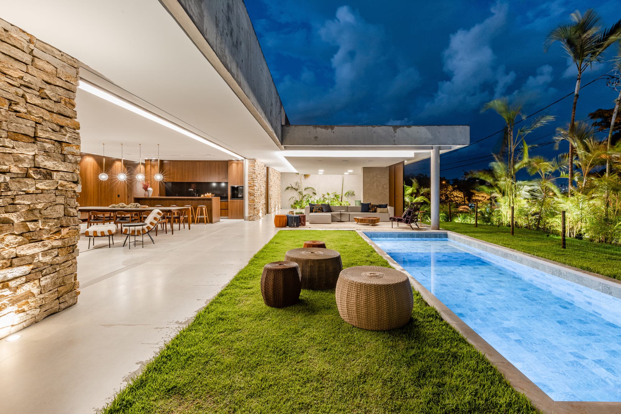 Área externa de casa com piscina linear, gramado, pufes de palha e varanda com forro de madeira